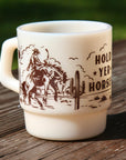An off-white ceramic mug with a brown western-themed graphic and the text 'HOLD YER HORSES' sitting on a weathered wooden surface with a blurred green outdoor background.