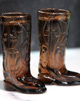 Pair of brown glass cowboy boot shot glasses on a white surface with a dark background