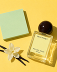 Whitney perfume bottle beside vanilla pods, a white flower and a mint green block on a yellow background