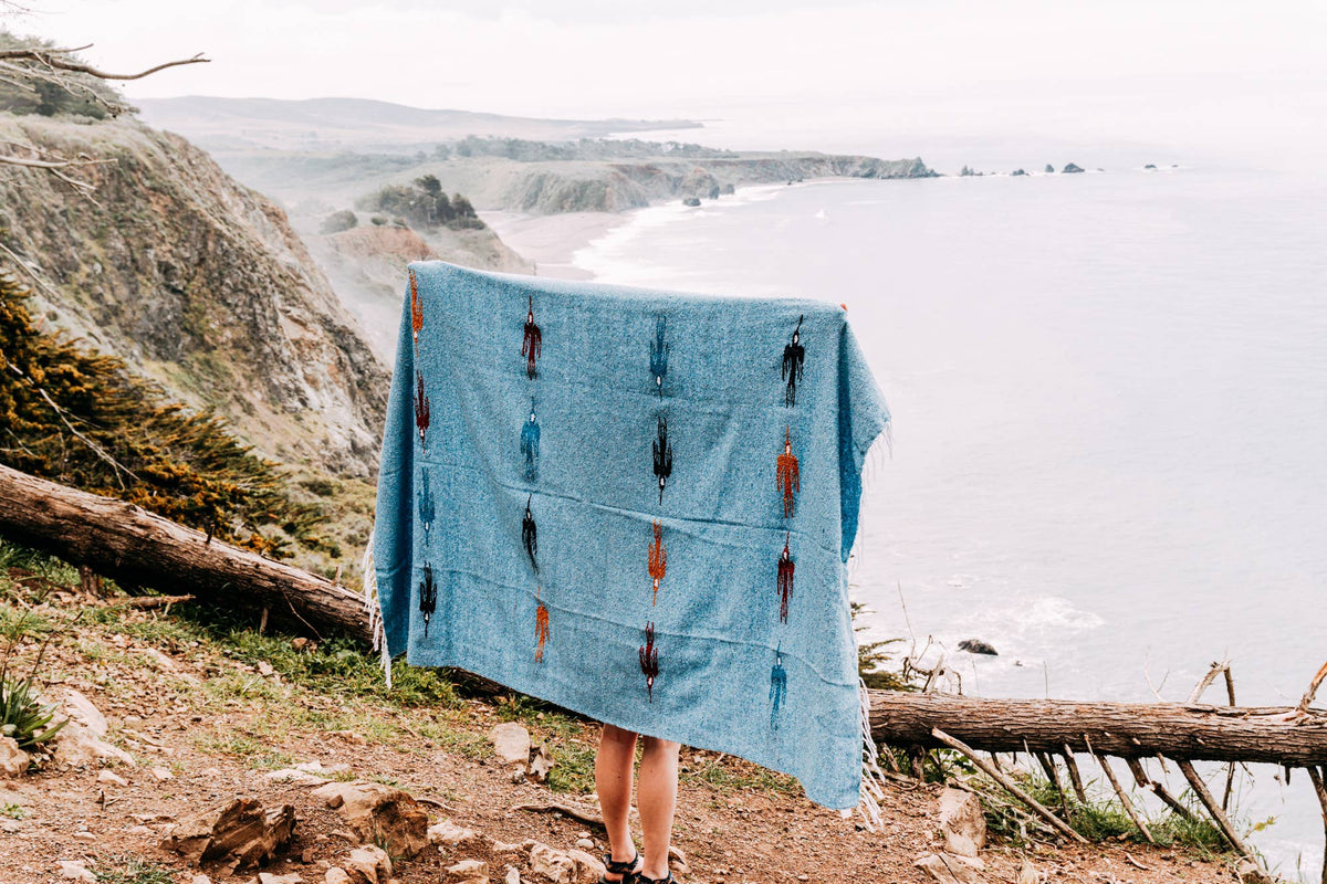 Person holding up a light blue thunderbird blanket on a cliff above the ocean