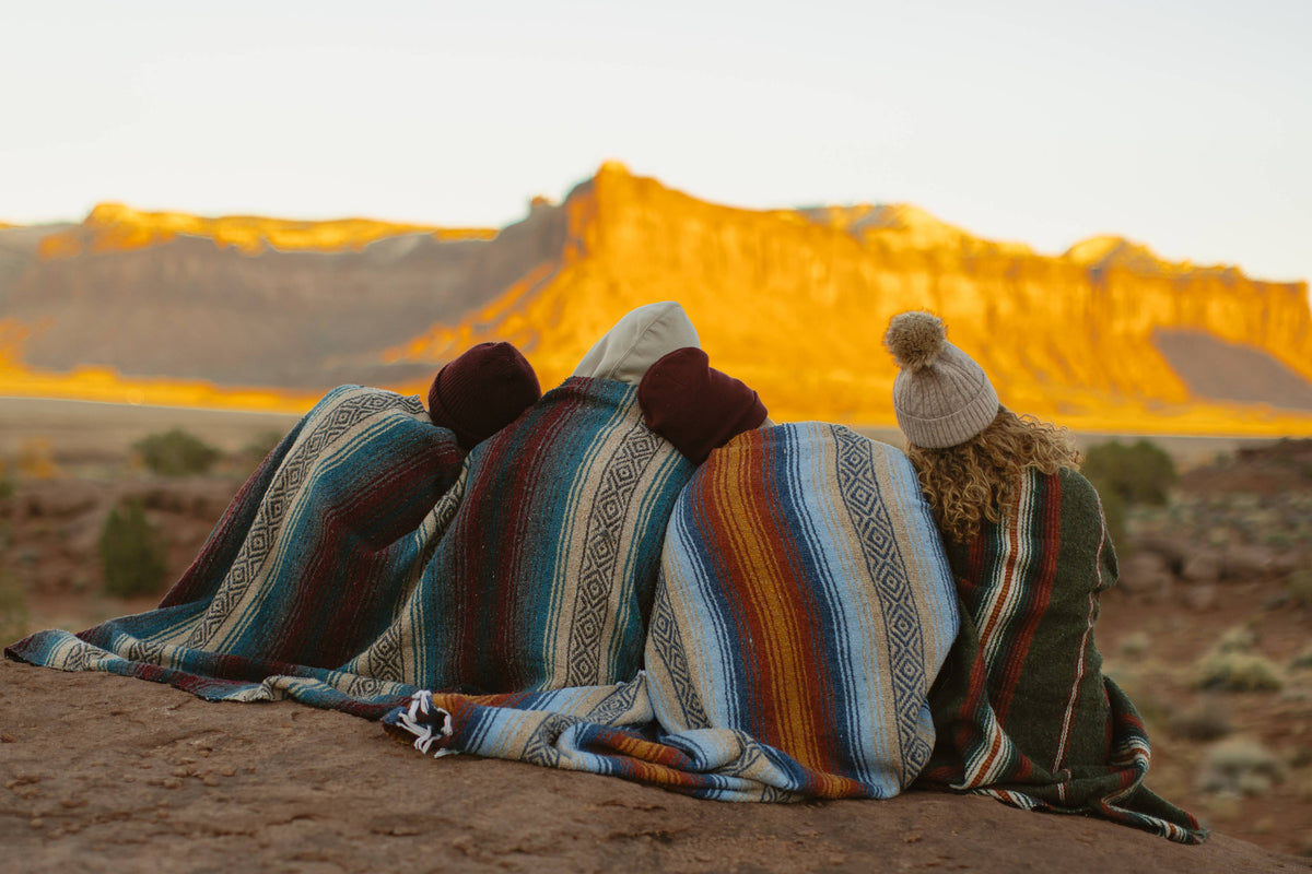 Three people wrapped in striped blankets sitting in the desert at sunset