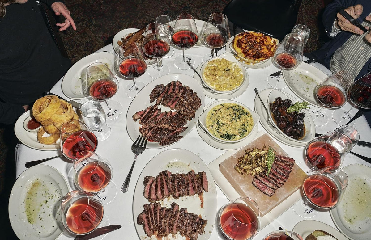 Overhead photo of a steakhouse table with sliced steaks, side dishes, onion rings and glasses of red wine