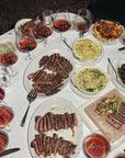 Overhead photo of a steakhouse table with sliced steaks, side dishes, onion rings and glasses of red wine