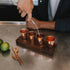 Person pouring a drink into four copper shot glasses on a walnut flight board with limes